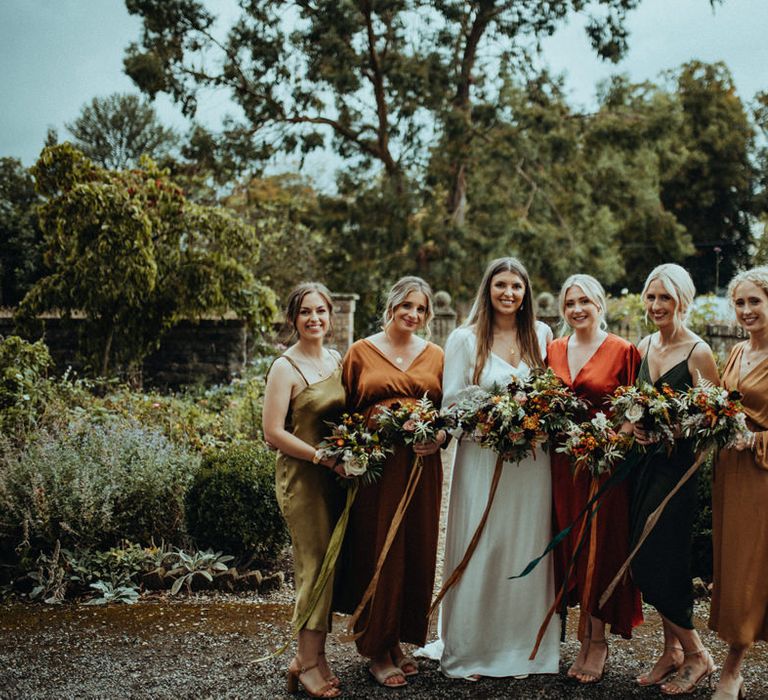 Bride stands with her bridesmaids who wear Autumnal coloured bridesmaids gowns in differing styles