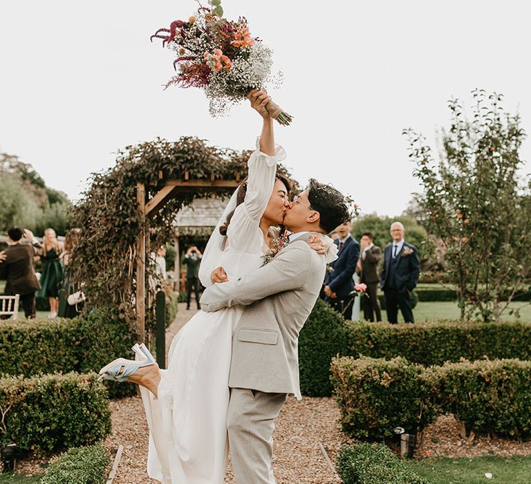 Groom lifts his bride as they kiss and bride holds up her floral bouquet for outdoor wedding