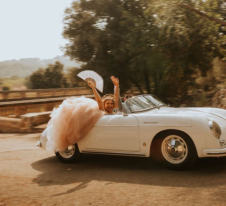 Bride & groom smile and wave as they drive in vintage car