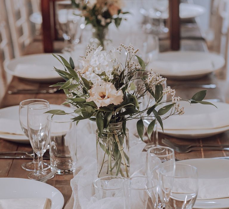 Pale pastel florals in clear vase next to white table runner on wooden table