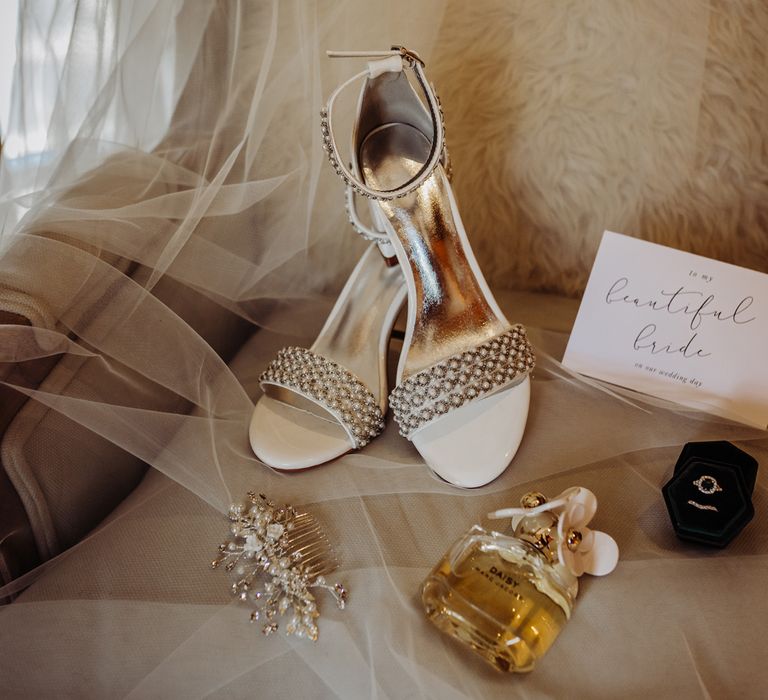 White heeled sandals with gold details gold and pearl hair clip, Daisy by Marc Jacobs perfume bottle, ring box and wedding stationery with modern calligraphy