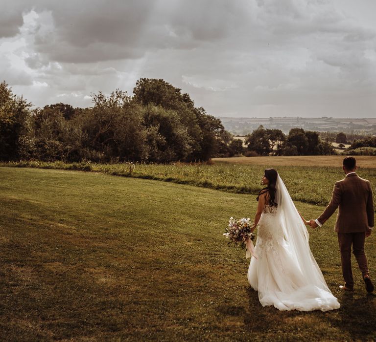 Bride in lace wedding dress with tulle train and veil holding multicoloured bridal bouquet walks holding hands with groom in brown tweed suit through the Cotswold countryside