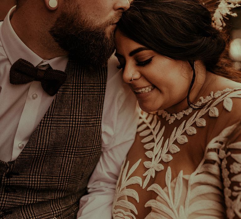 Golden hour portrait with groom in a tweed waistcoat and bow tie kissing his brides head in a boho lace wedding dress 