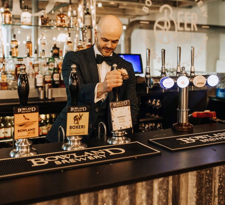 Groom in navy tweed suit and bow tie pours himself an ale at wedding reception