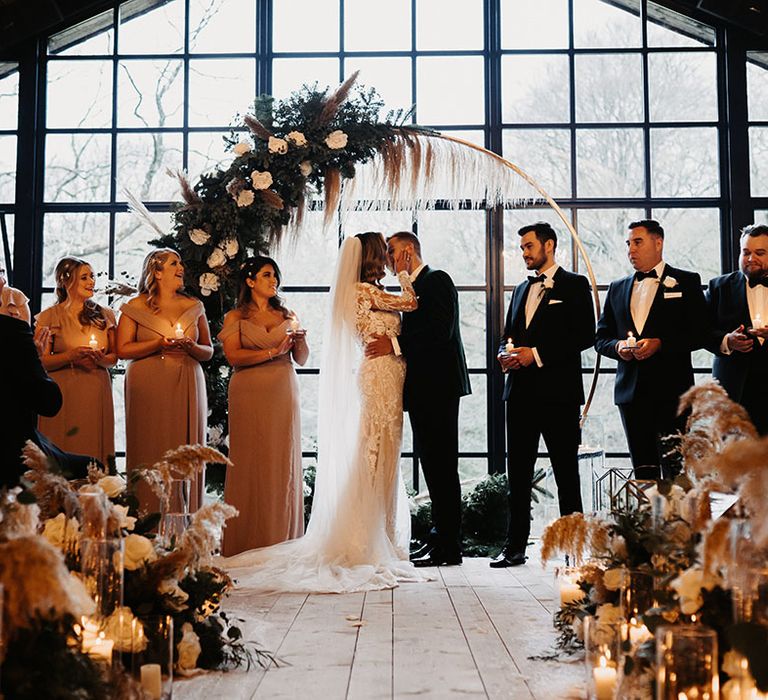 Bride and groom share a kiss after humanist wedding ceremony with bridal party and groomsman standing to the side holding candles 