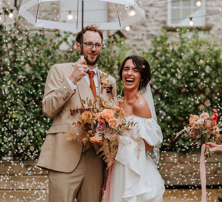 Bride in white puff sleeve wedding dress and Loeffler Randall shoes with groom in beige three piece suit holding clear umbrella as confetti is thrown 