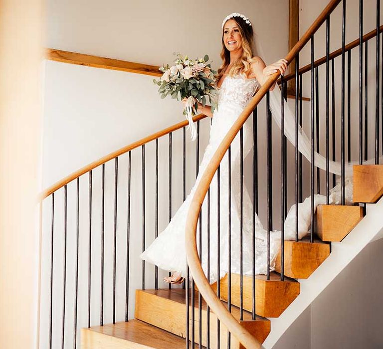 Bride walks down stairs in floral lace wedding dress with long veil and white headband holding rose bouquet 