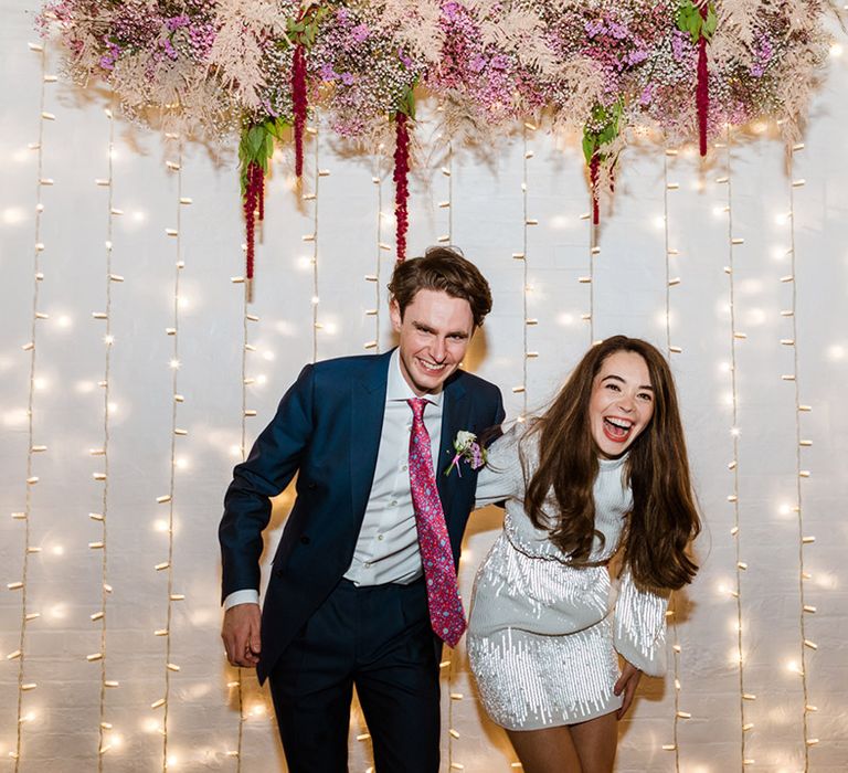 Bride and groom have a fun time at their party city wedding with bride in sparkly second outfit and groom in blue suit in front of fairy lights 