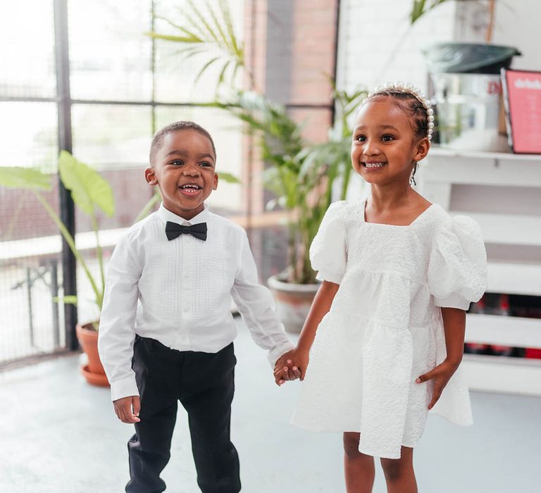 Page boy in black tie and white converse trainers holds hands with flower girl in white puff sleeve dress and pearl headband 