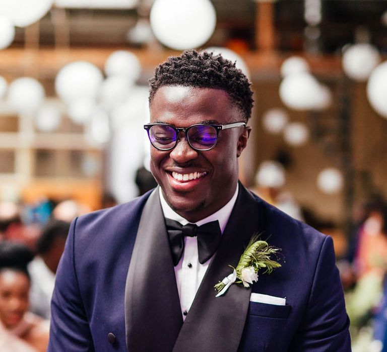 Groom in blue and black tuxedo and black glasses waiting at the altar for the bride with white flower buttonhole 