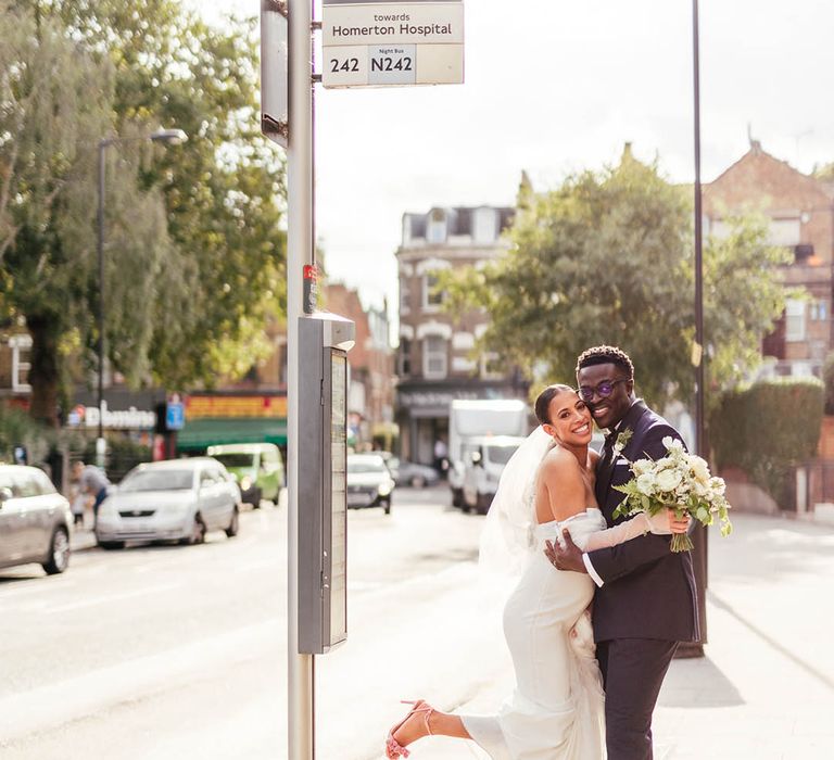 Groom in black tie and bride in off the shoulder corset top wedding dress with pink pom pom wedding shoes hug outside of a London bus stop