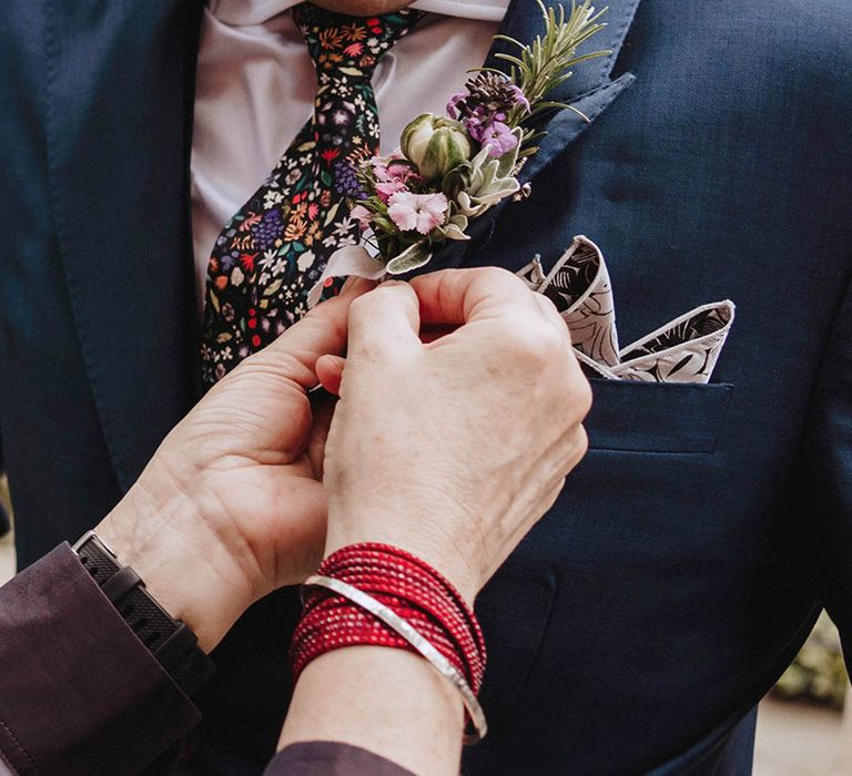 Groom in blue suit with floral tie and patterned handkerchief gets his buttonhole affixed to his jacket