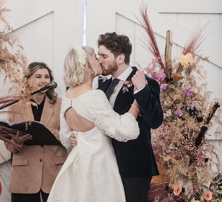 Bride in short wedding dress with platform black block heels kissing the groom in velvet suit next to dried wedding flowers