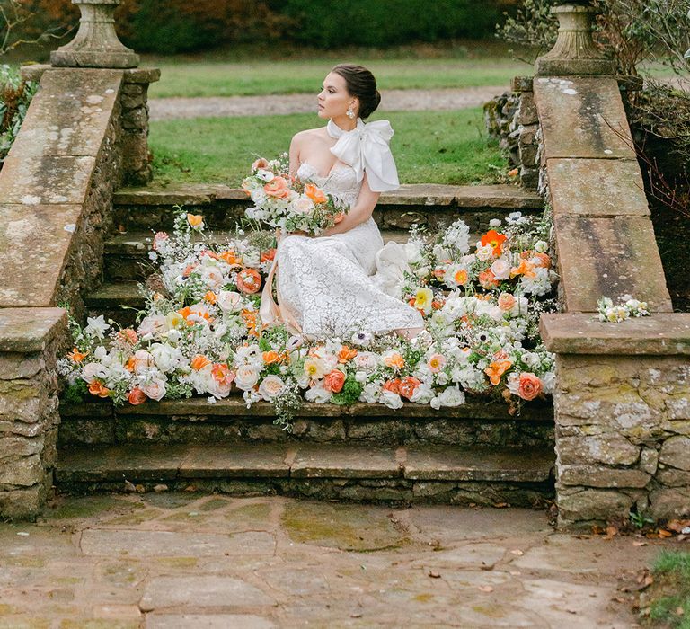 Bride with a pinned wedding up do in a strapless lace wedding dress with bow neck scarf sitting on the steps at Holesfoot surrounded by peach and white wedding flowers 