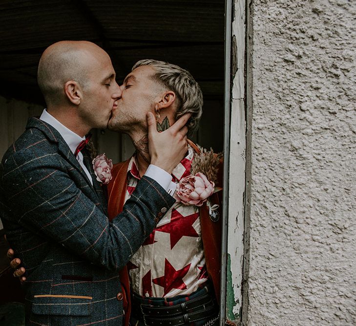 Groom wears colourful shirt and orange velvet blazer as he kisses his groom outdoors 