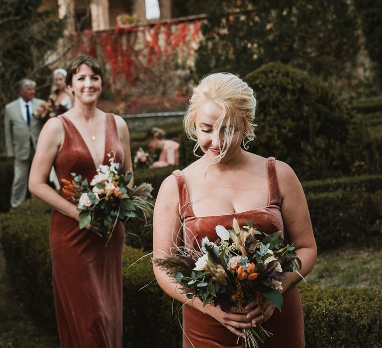 Bridesmaid walks down the aisle wearing velvet terracotta orange bridesmaids dresses finished with a square neckline and fitted style 