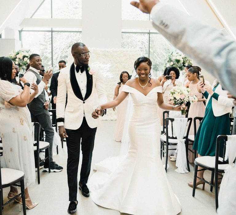 Groom wears suave white dinner jacket and black tie whilst holding his bride's hand wearing fitted off-the-shoulder wedding dress after ceremony