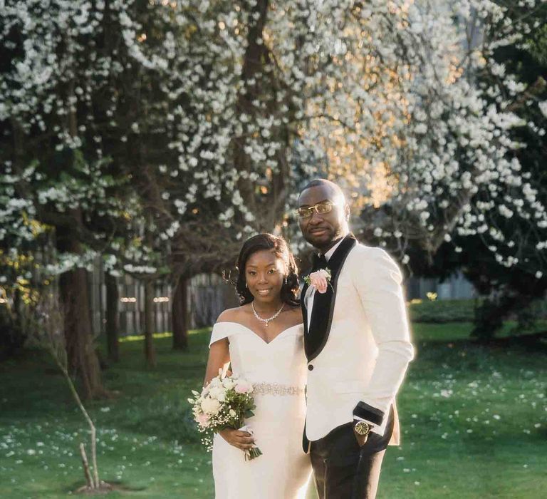 Bride wearing fitted wedding dress & groom in black tie stand outdoors on their wedding day for couples portraits 