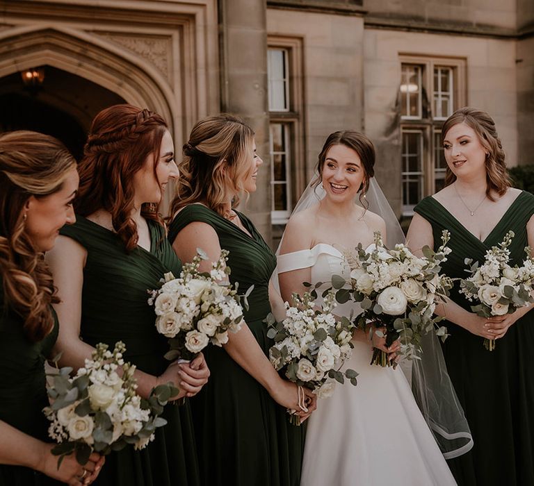 Bridesmaids in dark green dresses with the bride in an off the shoulder dress holding white flower bouquets 