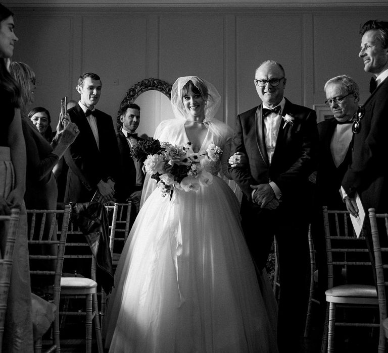 Bride wears hood styled cathedral veil and walks down the aisle with her father wearing black tie at Findon Place 