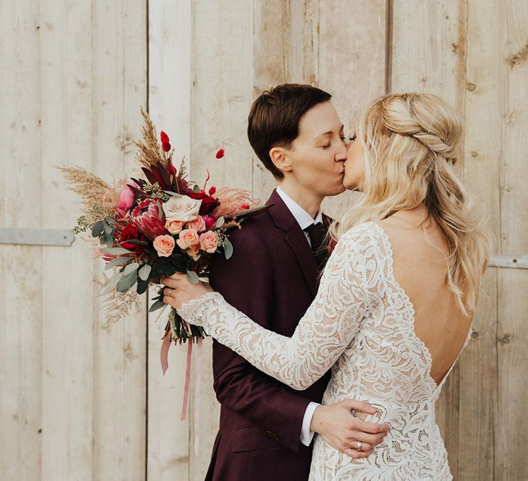 Bride in a long sleeve lace wedding dress with her blonde hair in a half up half down style with kissing her partner in a burgundy suit 