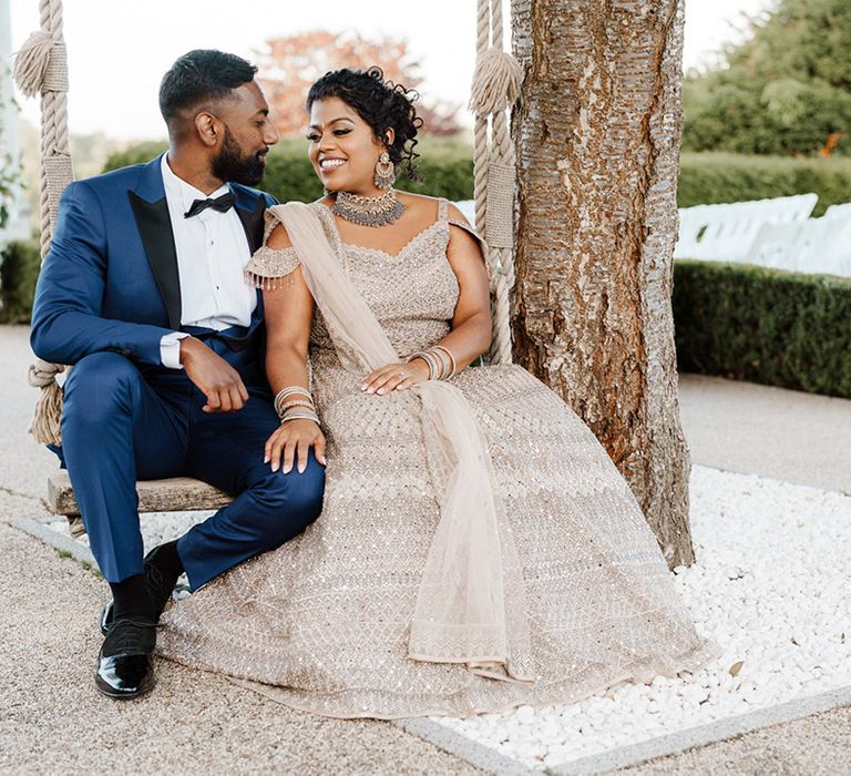 Bride in gold embellished dress sits beside her groom in black tie on outdoor swing 