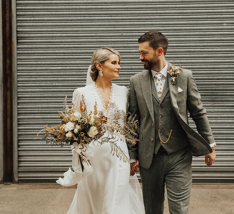 Bride in Reformation wedding dress with long sleeves walks with her groom in three-piece suit 