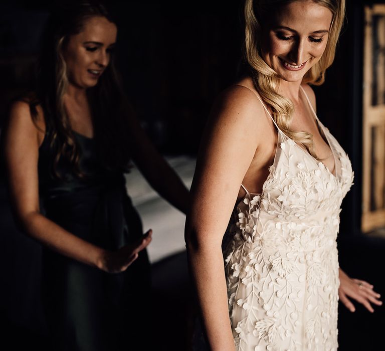 Bridesmaid in green satin dress helps the bride into her Alexandra Grecco Lana wedding dress with a pretty pearl headband 