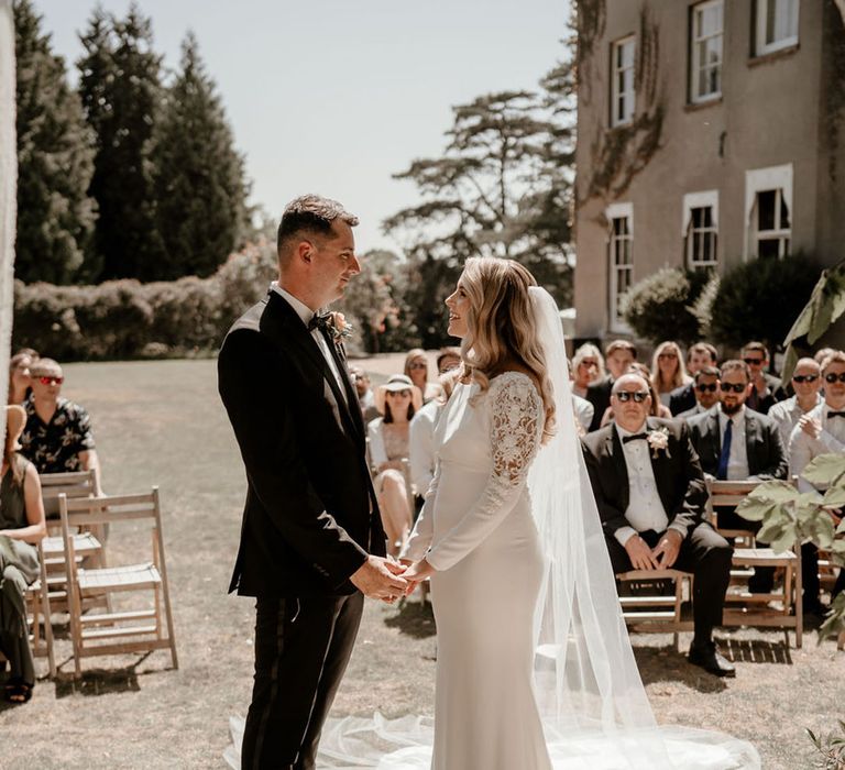 Bride with blonde hair smiling up at the groom as they hold hands at the altar for their outdoor wedding ceremony 