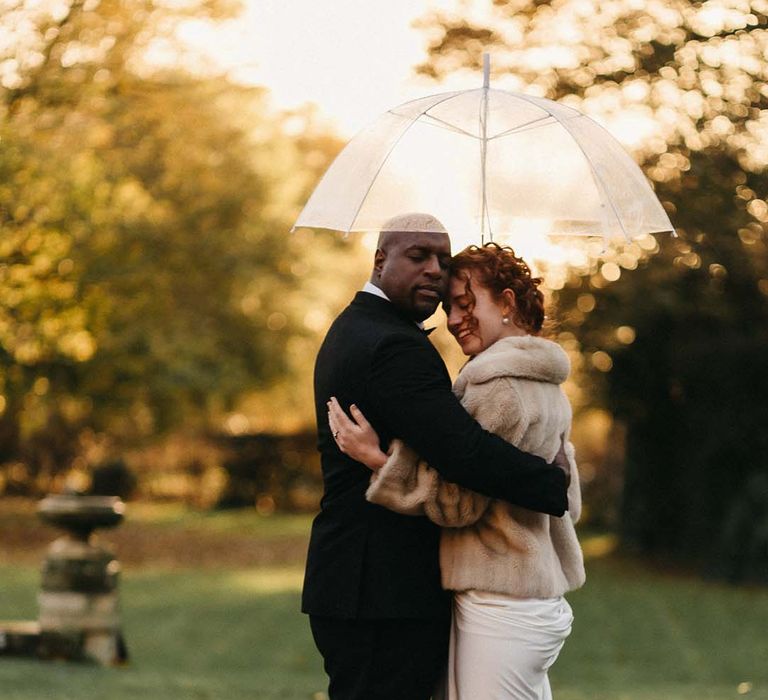 Groom in black tux embracing bride in satin white wedding gown, pearl hair accessories, white closed-toe heels and a light fur coat 