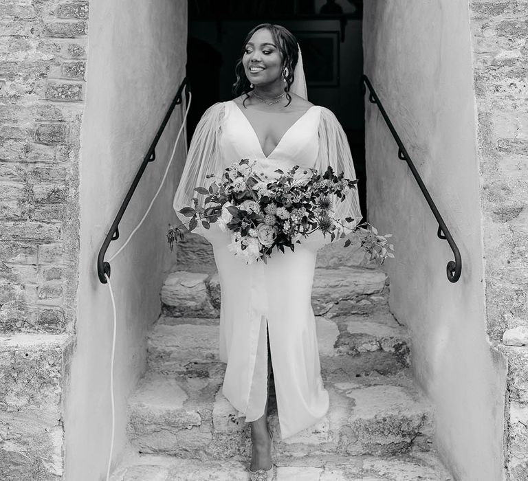 Bride walks down steps from archway of rustic Italian venue in her sheer-sleeved wedding dress, something blue wedding shoes and bouquet full of roses, wildflowers and thistles