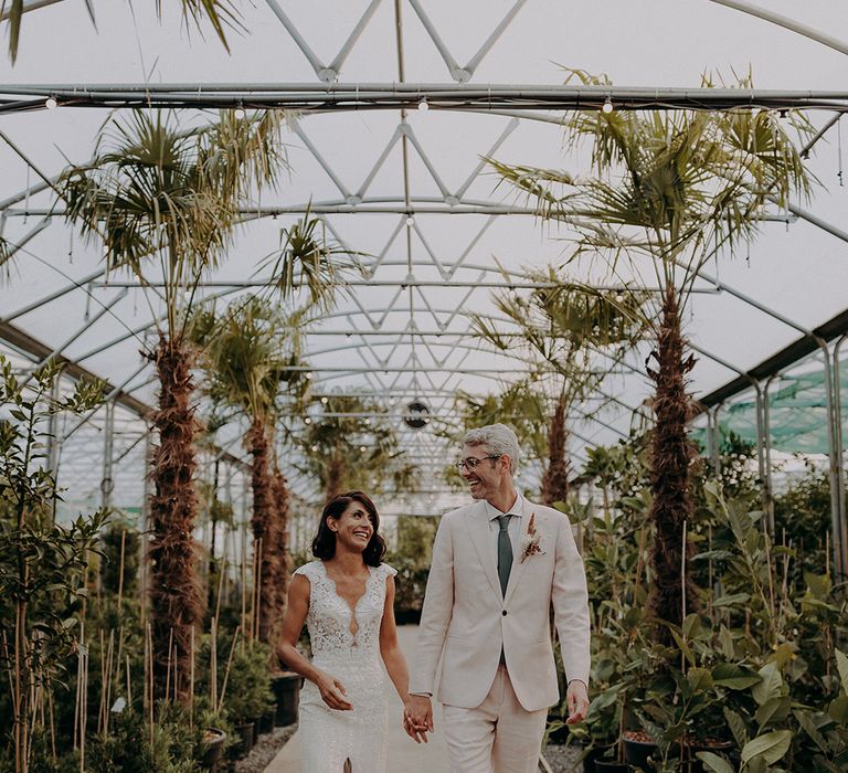 Bride in lace and tulle dress and groom in light pink suit hold hands whilst walking through plants and trees