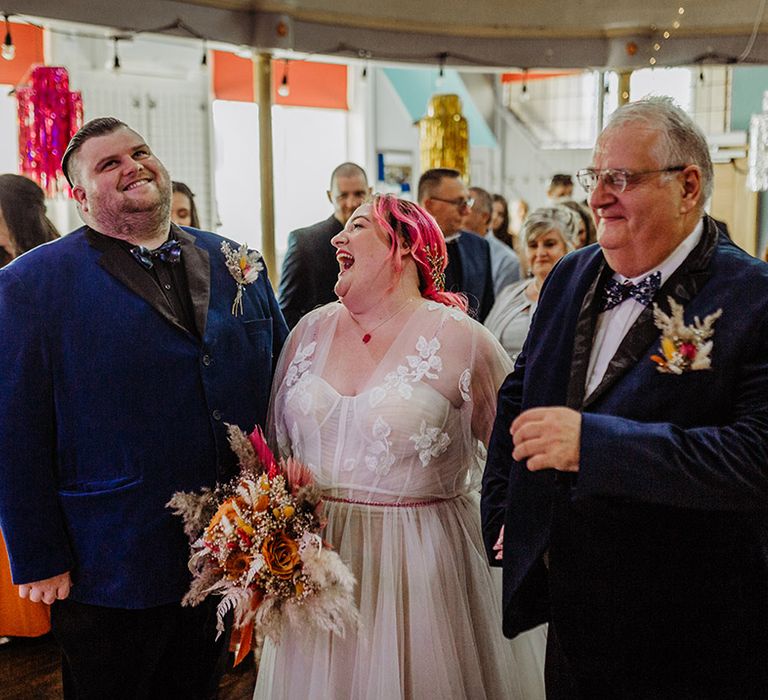 Bride in corset top wedding dress with leaf detailed lace overlay, pink stitching around the waist and red gemstone necklace holding autumnal coloured bouquet with white pampas grass and an orange ribbon and groom and father of the bride in midnight blue blazers with bowties and boutonnieres 