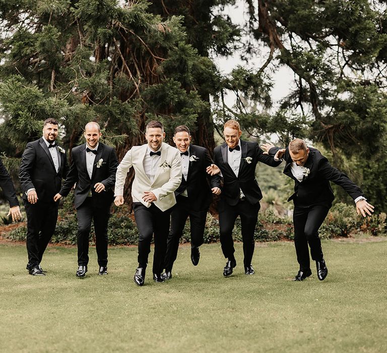 Groom in a white suit jacket and black trousers running with the groomsmen in black tie 