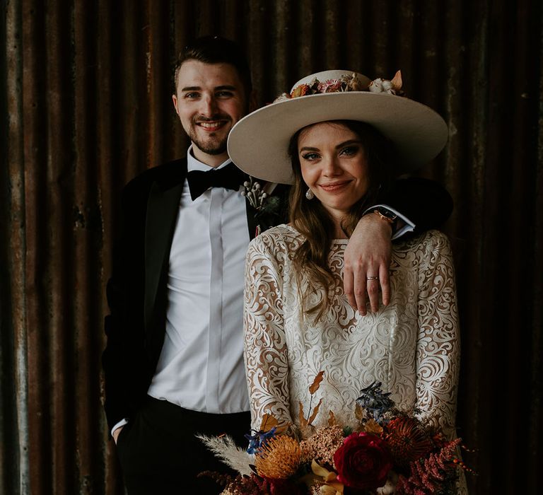 Groom in black tie rests his arm around the bride in a laser cut lace wedding dress with bell sleeves and white hat 