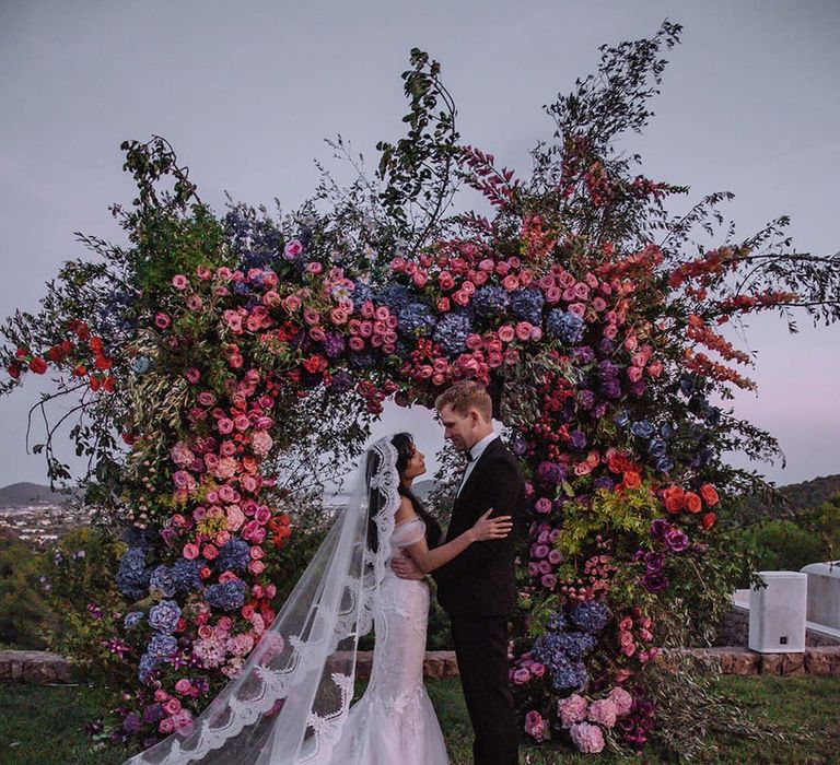 Bright and bold pink and blue wedding flower arch arrangement with the bride in a lace edge veil smiling at the groom 