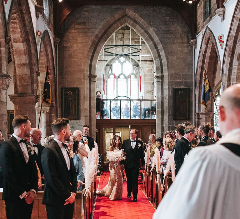 Father of the bride in a black tuxedo walks the bride down the aisle on a red carpet to the groom waiting at the end also in black tie 