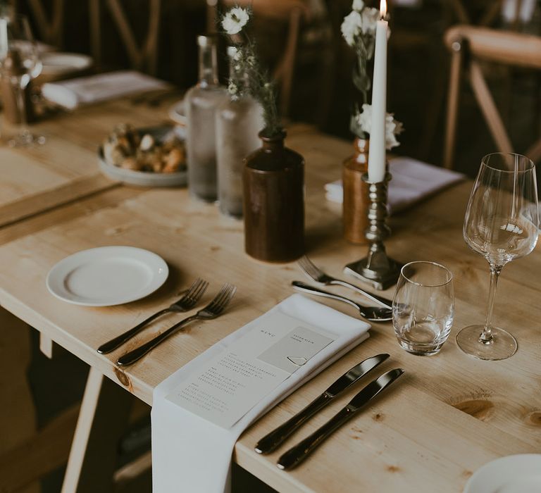 Rustic simple and elegant wedding tablescape with white taper candles, white napkin and wedding menus and small arrangements of flowers 