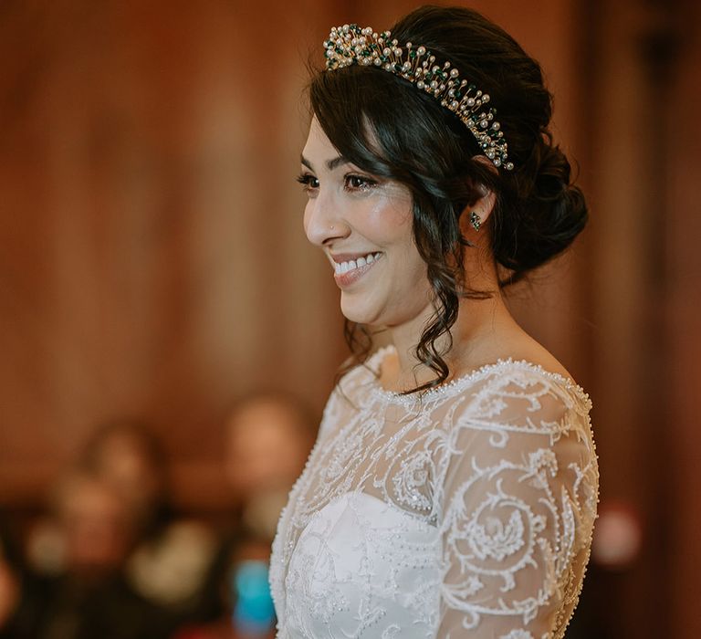 Bride in princess wedding dress with long lace sleeves and gold, pearl and emerald bridal crown standing at the alter