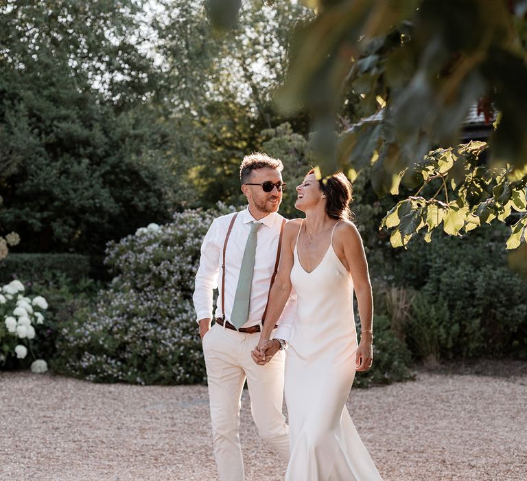 Bride in fitted silk and simple Grace Loves Lace wedding dress walks alongside her groom in sage green tie and braces