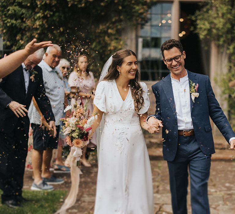 Bride with half up half down brown hair wearing a handmade embroidered wedding dress walking with the groom in a navy suit for their confetti exit 