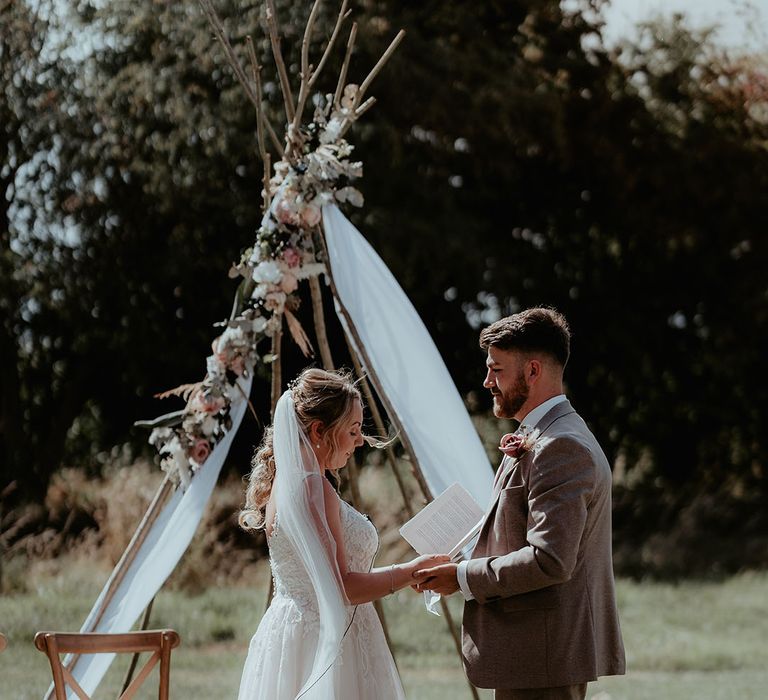 Bride & groom stand in front of DIY ceremony structure complete with wildflower decor and white drapes 