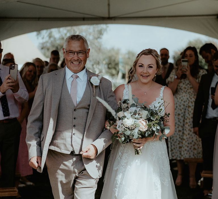 Bride walks down the aisle holding wildflower bridal bouquet alongside her father in neutral three piece suit and pink tie 