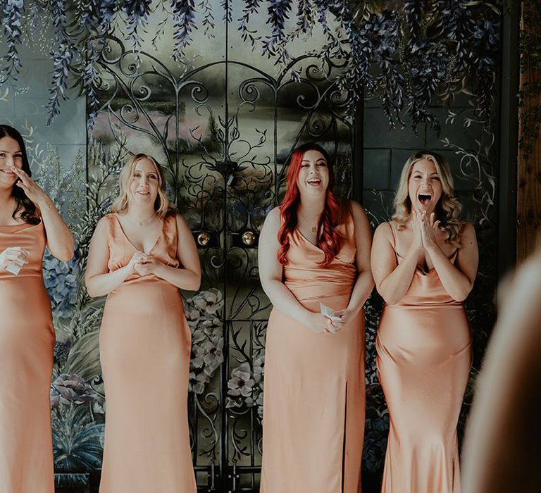 The bridesmaids in matching satin cowl neck peachy orange bridesmaid dresses smile as they get their first look at the bride 