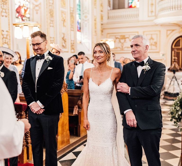 The bride wears a subtle mermaid fitted lace wedding dress from Grace Loves Lace standing with the groom and father of the bride in matching black tuxedos 