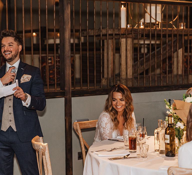 Groom in navy suit with spotted navy tie and cream waistcoat and pocket square reading out his wedding speech with a microphone with the bride smiling up at him