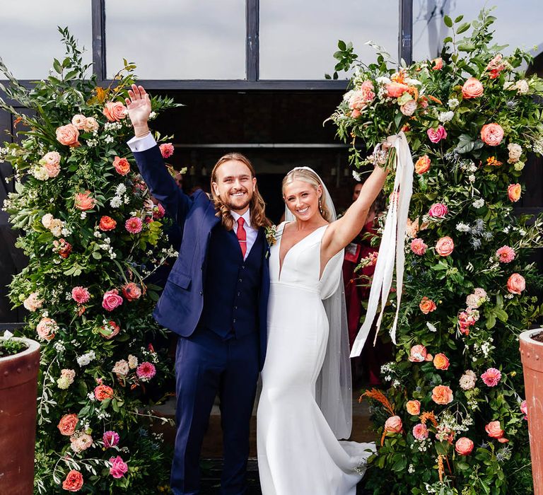 Bride in a fitted wedding dress with pearl earrings with the groom in blue three piece suit celebrating with the bride next to the flower column arrangements