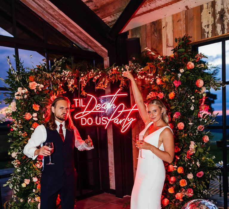 The bride and groom pose next to their red neon wedding sign reading 'til death do us party' with flower column decor 