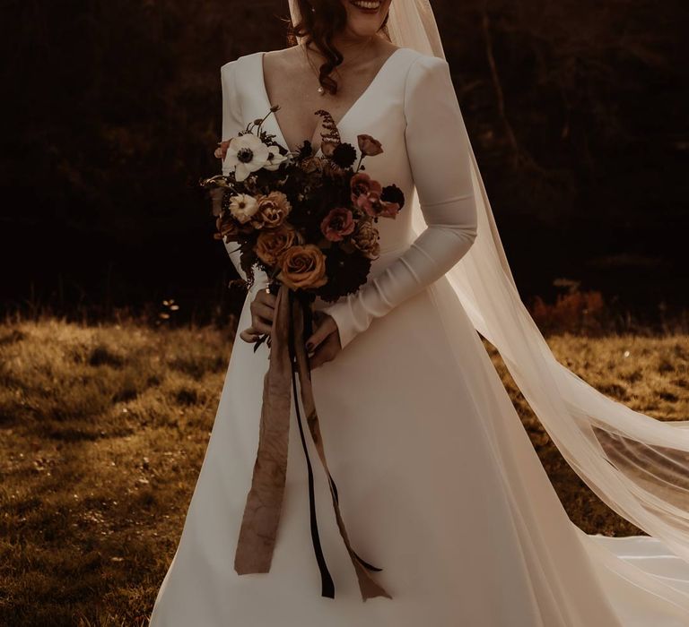 Bride in long sleeve deep v wedding dress and church length veil holding white, dusky pink and burnt red dried flower, rose and peony bridal bouquet at Hidden River Cabins 