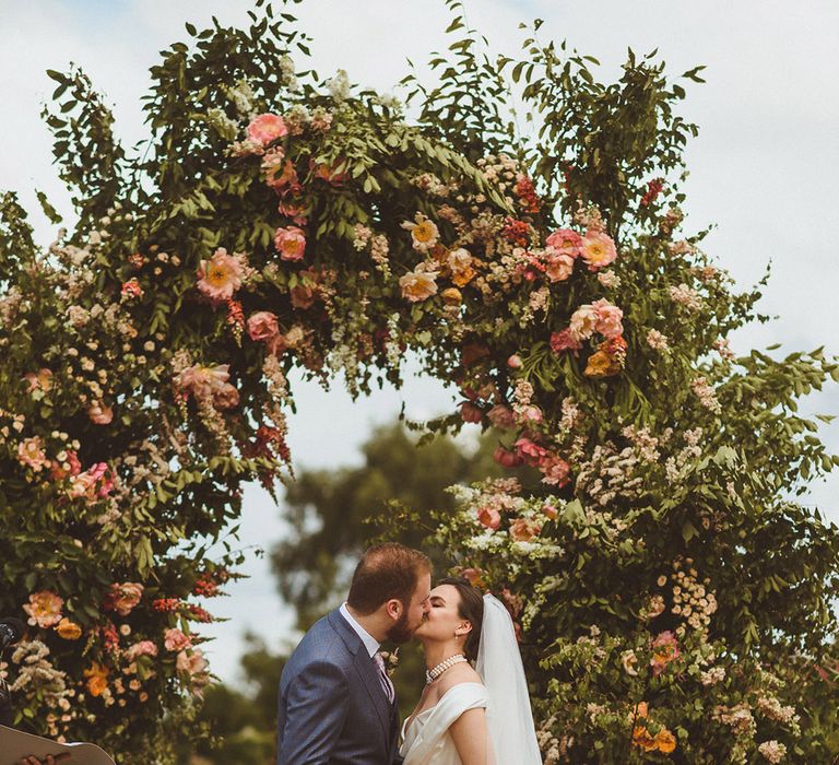 Bride wearing a pearl choker necklace by Vivienne Westwood with the groom in blue suit in front of floral arch altar decoration 